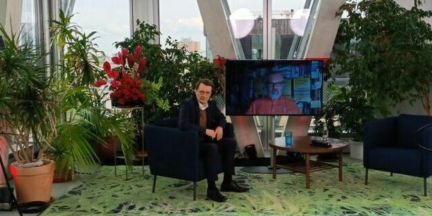 Karl Lauterbach sitzt auf einem Lounge-Stuhl zwischen Pflanzen, neben ihm ein Bildschirm, dort ist Jan Feddersen zu sehen Karl Lauterbach sitzt auf einem Lounge-Stuhl zwischen Pflanzen, neben ihm ein Bildschirm, dort ist Jan Feddersen zu sehen