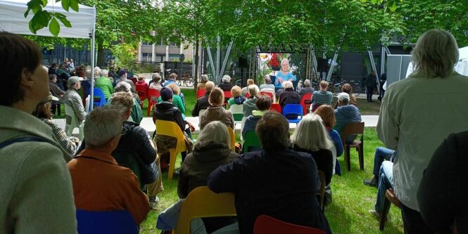 viele Menschen sitzen im Park auf bunten Stühlen und schauen auf einen Bildschirm