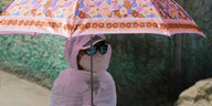 Eine Frau komplett verhüllt in weißen Stoff, trägt einen Sonnenschirm, eine FFP2-Maske und eine Sonnenbrille