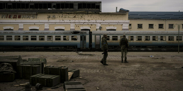 Zweiuniformierte Männer stehen im Vordergrund an einem Bahnsteig. Im Hintergrund steiht ein zerstörter Zug mit zerborsten Scheiben. Das Bahnhofsgebäude dahinter hat unzählige Einschusslöcher Zweiuniformierte Männer stehen im Vordergrund an einem Bahnsteig. Im Hintergrund steiht ein zerstörter Zug mit zerborsten Scheiben. Das Bahnhofsgebäude dahinter hat unzählige Einschusslöcher