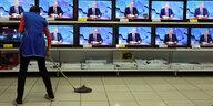 Eine Frau mit einer blauen Schürze wischt einen hellen Fliesenboden. Im Hintergrund sieht man ein mit Fernsehern gefülltes Regal. Auf allen Bildschirmen ist der gleiche Mann zu sehen. Es ist der russische Präsident Wladimir Putin.