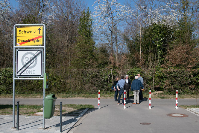 Eine Gruppe von Leuten steht nahe einem Ortsschild das den Weg in die Schweiz anzeigt