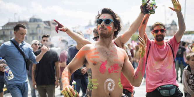 Ein oberkörperfreier, geschminkter Mann mit Sonnenbrille trägt eine Sonnenbrille und hält ein Bier in seiner Hand.