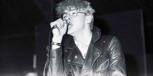 Schwarz-weiß Aufnahme: Der Musiker Alexander Hacke steht in Lederjacke auf der Bühne und singt in ein Mikrofon. Er trägt eine Brille.