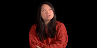 Eine Frau im roten Pullover vor einem schwarzen Hintergrund erzählt