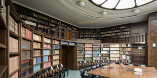 In einem Raum stehen in einem Halbrund hölzerne Regale voller Bücher. Davor stehen ein Tisch und Stühle, an der Decke ist ein rundes Overlicht