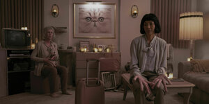 Jamie Lee Curtis und Michelle Yeoh sitzen mit Wurstfingern in einem Wohnzimmer.