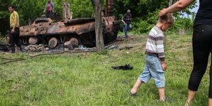 Eine kleiner Junge wird von einer Frau am Kopf gestrichen, sie führt ihn von einem ausgebrannten Panzer weg