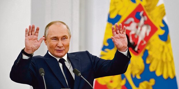 Präsident Putin mit russischer Flagge im Hintergrund hebt die Hände hoch