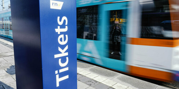 Ein blauer Ticketautomat an einer Haltestelle, im Hintergrund eine fahrende Straßenbahn