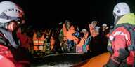 Aus Seenot gerette Menschen sitzen mit Schwimmwesten auf einem Schlauchoot, Helfer*innen davor