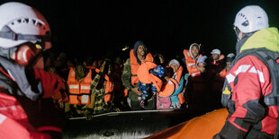 Aus Seenot gerette Menschen sitzen mit Schwimmwesten auf einem Schlauchoot, Helfer*innen davor