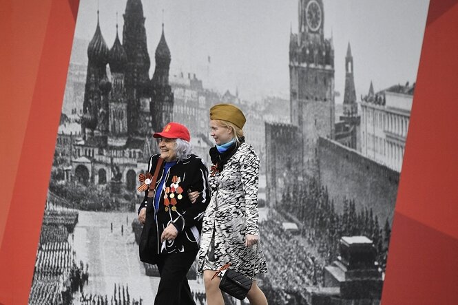 Eine junge und eine alte Frau laufen vor einem Plakat mit dem historischen Foto von einer Militärparade auf dem Roten Platz