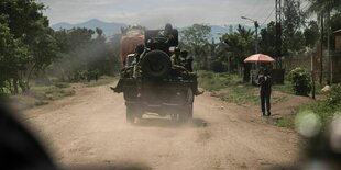 Ein Panzer beladen mit Soldaten fährt auf ener staubigen Straße, auf der ein Mann mit pinkfarbenenem Sonnenschirm geht