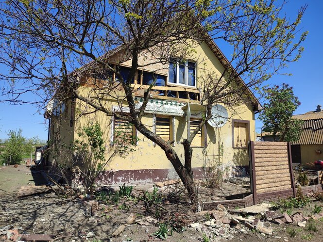 Ein gelbes Haus, davor ein Baum, die Erde ringsum sieht nicht gepflegt aus, aber es sprießen ein paar Pflanzen davor. Der Himmel ist strahlend blau