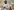Der Rapper Akon steht in einer Moschee und Betet. Er trägt ein gediegenes Hemd in Grauton und hat die Hände unter der Brust übereinandergeschlagen. Er trägt eine Maske. Im Hintergrund weitere Männer.