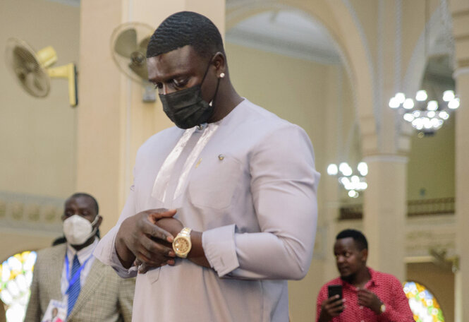 Der Rapper Akon steht in einer Moschee und Betet. Er trägt ein gediegenes Hemd in Grauton und hat die Hände unter der Brust übereinandergeschlagen. Er trägt eine Maske. Im Hintergrund weitere Männer.