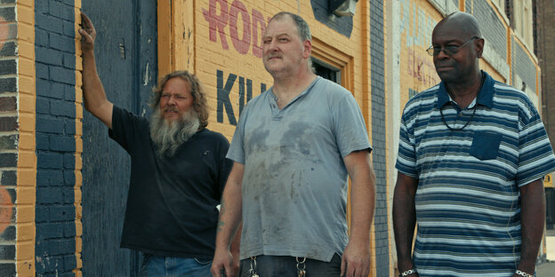 Drei Männer, zwei weiß, einer schwarz, stehen in einem Gewerbegebiet auf der Straße.