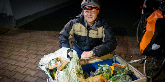 Ein Mann mit Beleuchtung am Fahrradhelm steht über einem Fahrradanhänger, der mit Lebensmitteln gefüllt ist