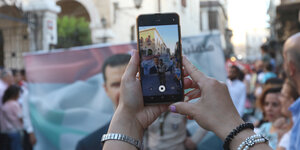 Zwei Hände im Vordergrund des Bildes halten ein Smartphone, das mit der Kamera einen Soldaten fokussiert, der bei einer Demonstration vor einem Banner mit einem Mann darauf steht. Der Mann auf dem Banner is Baschar al-Assad, der Präsident Syriens.