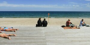 Menschen sitzen bei Sonnenschein am Rand eines leeren Strands und niemand im Wasser, ein Schild weist auf die Minengefahr hin