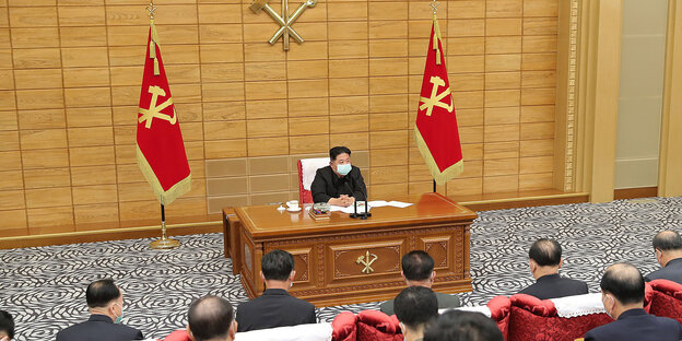 Diktator Kim Jong Un sitzt mit Mundschutz alleine hinter einem Pult auf einer Bühne, flankrt von zwei Nationlflaggen