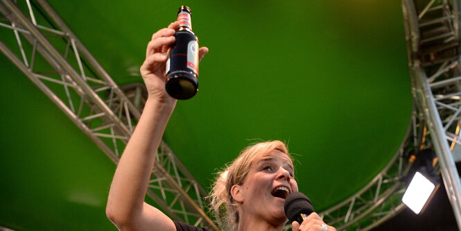 Mona Neubaur hebt die Bierflasche vor grüner Wand