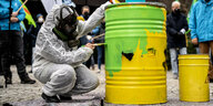 Ein Mensch mit Gasmaske und Schutzanzug malt ein symbolisches gelbes Atomfass in grün an