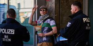 Ein Mann macht das Victory Zeichen, er trägt eine Broschüre für Palästina in der Hand und steht zwischen 2 Polizisten