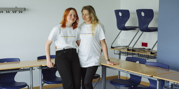 A mother and daughter are standing side by side in a classroom, both wearing white t-shirts and black pants