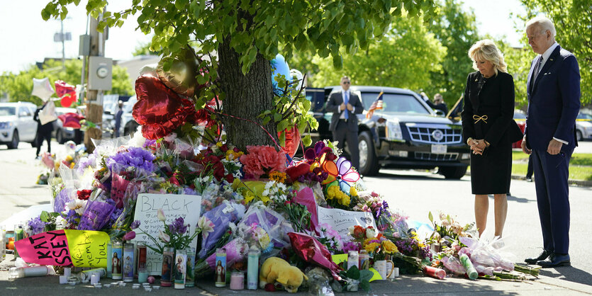 Um einen Baum sind unzählige Blumen aufgetürmt. Joe und Jill Biden stehen andächtig davor