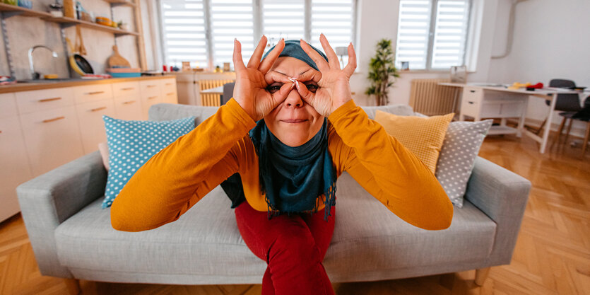 Junge Frau mit kopftuch hält sich die Hände vor die Augen wie ein Fernglas und sitzt auf einem Sofa