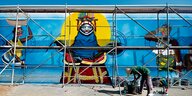 Ein Maler beugt sich über Eimer, hinter ihm eine blaue Wand mit Figuren und ein Gerüst zum Arbeiten