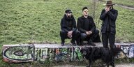 Szene im Freien: Die drei Mitglieder der Band Sudden Infant sitzen und stehen auf einer Mauer in einem Park, sie sind schwarz angezogen. Vor ihnen steht ein Hund