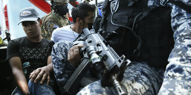 Zwei Männer in Zivil sitzen neben bewaffneten Soldaten