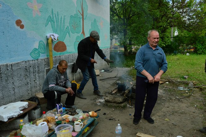 Drei alte Männer rund um die beschriebene Feuerstelle. Die Hauswand ist türkis bemalt, neben den Männern stehen auf Paletten ein paar Lebensmittel und Becher. Äpfel in Plakstiktüten u.ä.