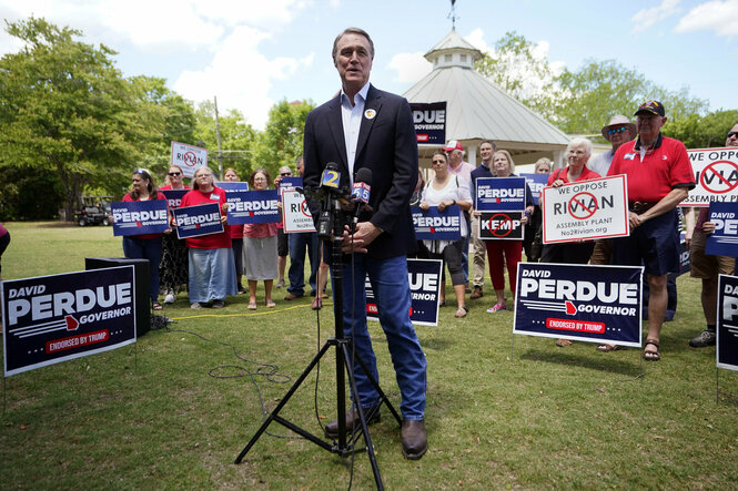 Ein Mann im dunklen Jacket spricht in ein Mikro, hinter ihm auf dem Rasen Menschen mit Schildern "David Perdue"