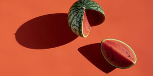 Auf rotem Hintergrund liegt eine Wassermelone, von der etwa ein Viertel abgeschnitten ist. Das abgeschnittene Stück liegt neben der restlichen Melone.