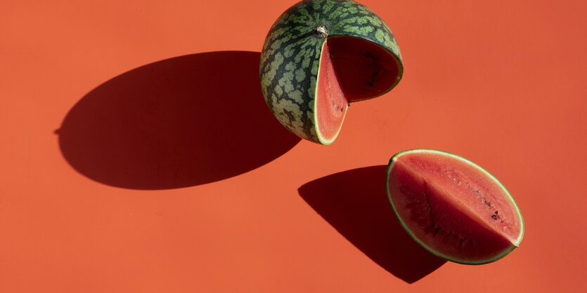 Auf rotem Hintergrund liegt eine Wassermelone, von der etwa ein Viertel abgeschnitten ist. Das abgeschnittene Stück liegt neben der restlichen Melone.