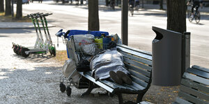 Scheinbar obdachloser Mensch liegt auf einer Bank im Schatten eines Baumes.