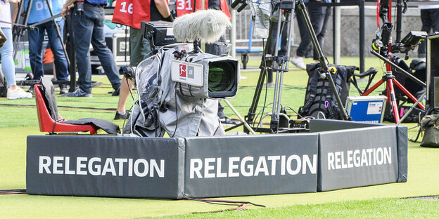 Eine Fernsehkamera am Rande eines Fußballspielfeldes. Auf Schildern davor steht das Wort "Relegation"