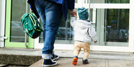 Eine Erwachsene Person läuft mit einem sehr jungen Kind an der rechten Hand und einem Rucksack in der Linken Hand auf eine Tür zu.