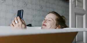 Eine Frau liegt in der Badewann und schaut auf ihr Handy, das sie in der Hand hält.