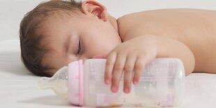 Ein Baby mit nacktem Oberkörper liegt auf dem Bauch. Es hat kurze dunkle Haare und seine linke Hand umklammert eine Flasche.