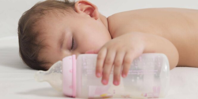 Ein Baby mit nacktem Oberkörper liegt auf dem Bauch. Es hat kurze dunkle Haare und seine linke Hand umklammert eine Flasche.