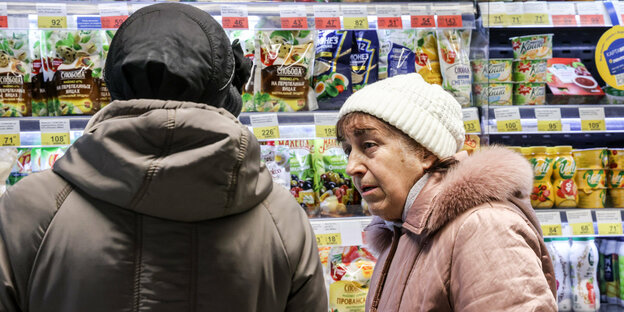 Zwei ältere Frauen in dicken Jacken stehen vor einem Lebensmittelregal. Die Beschriftung auf den Verpackungen ist in Kyrillisch.
