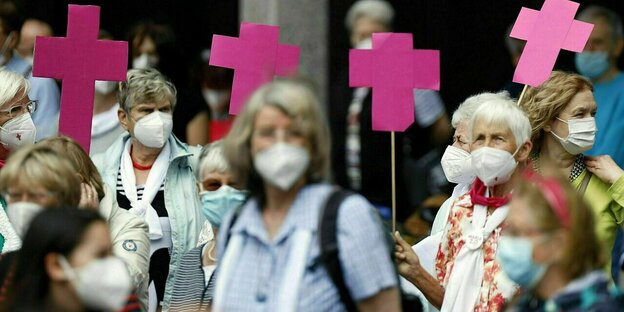 Frauen tragen Masken und protestieren mit pinkfarbenen Kreuzen gegen die frauenfeindliche Kirche