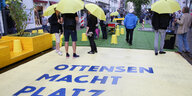 Menschen mit Regenschirmen auf einem markierten Stück Straße, auf dem "Ottensen macht Platz" steht