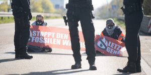 Zwei Aktivist:innen der Gruppe „Aufstand der letzten Generation“ blockieren mit Plakaten die Straße, vor ihnen stehen bewaffnete Polizist:innen