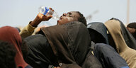 Flüchtlinge mit verhülltem Gesicht stehen in einer Reihe, einer reckt den Kopf und trinkt Wasser aus einer Flasche